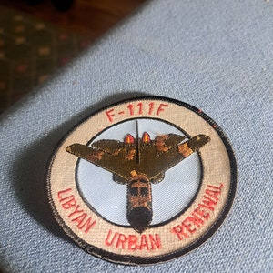 May include: A round, tan and black embroidered patch with a white circle in the centre. The circle features a brown and green F-111F fighter jet with red flames. The text "LIBYAN URBAN RENEWAL" is written in red around the circle.