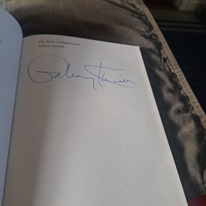 May include: An open book showing the title "The Book of Nightmares" by Galway Kinnell. A blue ink signature is visible on the white page. The text is in black.