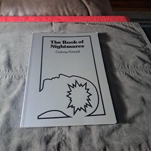 May include: A silver book titled "The Book of Nightmares" by Galway Kinnell. The cover features a black line drawing of a face and a starburst shape. The book is on a grey surface.