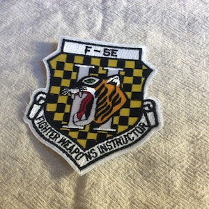 May include: White embroidered patch with a black and yellow checkered background. A tiger head with green eyes is in the center of the patch. The text "F-5E Fighter Weapons Instructor" is around the edge of the patch.
