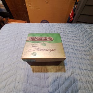 Könnte beinhalten: Eine Vintage View-Master Stereoskop-Box. Die Box hat eine gold-braune Basis mit einem grünen und weißen Deckel. Auf der Oberseite der Box stehen die Wörter "View-Master" und "Stereoskop".