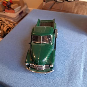 May include: A dark green die-cast model pickup truck featuring a chrome front grille, silver-toned headlights, a windscreen with two wipers, and a spare tyre mounted on the side. The vehicle has a classic cab design and an open truck bed.