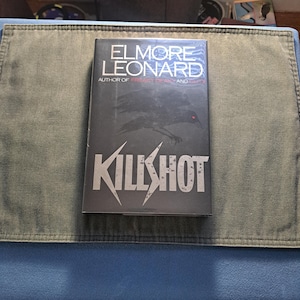 Puede incluir: Un libro de tapa dura titulado "Killshot" de Elmore Leonard. La portada es predominantemente gris oscuro con una ilustración estilizada de un cuervo y el título en letras blancas grandes y angulares. El nombre del autor está en blanco en la parte superior.