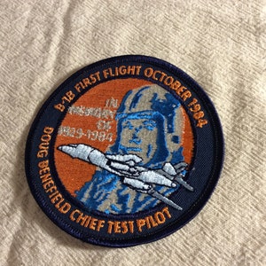 May include: A blue and orange embroidered patch with a pilot in a helmet and a plane. The text reads "B-1B First Flight October 1984 In Memory Of 1929-1984 Doug Benefield Chief Test Pilot".