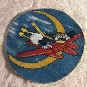 May include: A circular patch featuring a cartoon duck in a pilot's cap and goggles, riding a red airplane. The duck is set against a blue background with a yellow crescent moon and white clouds. The patch has a distressed look.