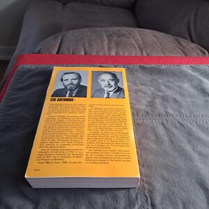 May include: A vintage book with a yellow cover featuring two black and white portraits of men. The title and author information are printed in black text. The book is resting on a gray blanket.