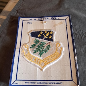 Puede incluir: Parche bordado con diseño de escudo. El parche presenta un escudo azul y dorado con el texto "EAST AIR DIVISION". El texto "N. S. MEYER, INC." está impreso en la parte superior. El texto "OUR SHIELD GUARANTEES DEPENDABILITY" está impreso en la parte inferior. El año "1966" está impreso en la parte inferior.