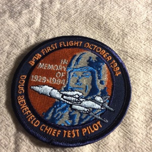May include: A blue and orange embroidered patch with the text "B-1B First Flight October 1984 In Memory Of 1929-1984 Doug Benefield Chief Test Pilot". The patch features a stylized image of a pilot in a helmet and a jet aircraft.