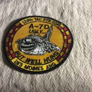 May include: A yellow and red embroidered patch with a cartoon A-7D aircraft and the text "124th TAC FTR SOB A-7D UGLY... BUT WELL HUNG DES MOINES ANG".