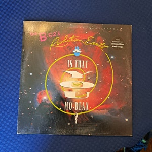 May include: A vinyl record album cover for The B-52's "Revolution Earth" single. The cover features a red and black cosmic design with the text "IS THAT MO-DEAN" in white. The B-52's and Revolution Earth are in pink and yellow.