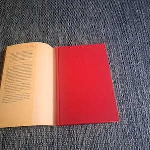 May include: An open book with text on the left page and a red hardcover book on the right. The red book has the initials "GB" embossed on the cover. The book is lying on a blue denim surface.