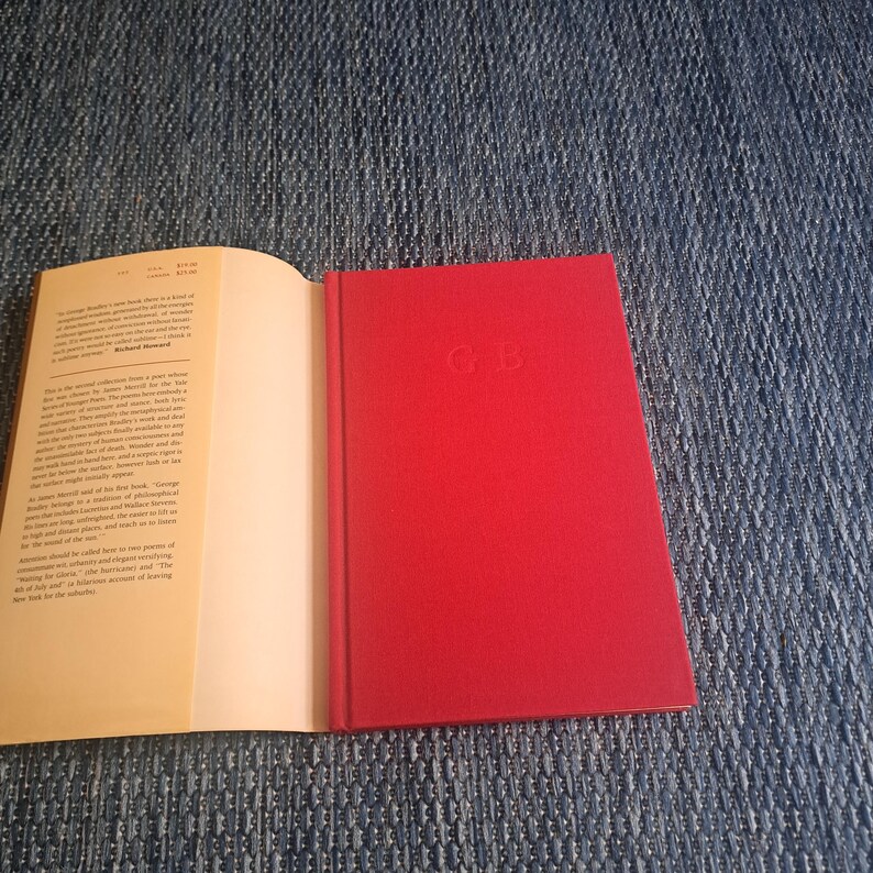 May include: An open book with text on the left page and a red hardcover book on the right. The red book has the initials "GB" embossed on the cover. The book is lying on a blue denim surface.