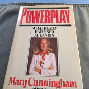 May include: A book titled "POWERPLAY" with the subtitle "WHAT REALLY HAPPENED AT BENDIX." The cover features a photo of Mary Cunningham and the author's name in red. The book is on a blue denim surface.