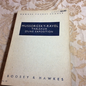 May include: A white book with blue text on the cover. The title is "Mussorgsky-Ravel Tableaux D'Une Exposition". The book is published by Boosey & Hawkes and is number 32 in their series. The price is $3.50.