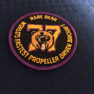 May include: A purple and gold embroidered patch with a bear and the text "Rare Bear" and "World's Fastest Propeller Driven Aircraft".