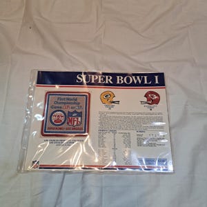 May include: A commemorative Super Bowl I display sheet featuring a square embroidered patch with the text First World Championship Game AFL vs NFL, team helmets for the Green Bay Packers and Kansas City Chiefs, starting lineups, and scoring.