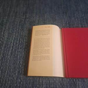 May include: An open book with a red cover and white pages. The right side of the book is open, revealing text. The cover has the initials "GB" in the lower right corner. The book is on a blue denim surface.