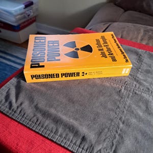 May include: A hardcover book titled "POISONED POWER" with a yellow cover and a black and orange radiation symbol. The book is by John W. Gofman and Arthur R. Tamplin. The book is resting on a gray fabric.
