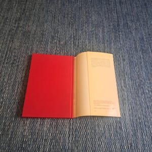 May include: An open book with a bright red cover and cream-colored pages. The book is lying on a blue denim surface. The right page contains printed text.