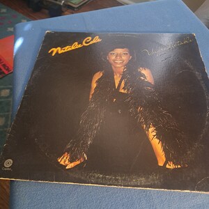 May include: A black vinyl record album cover featuring Natalie Cole. The album title is "Unforgettable" and is written in a cursive font. The album cover features a photo of Natalie Cole wearing a black dress and a feathered boa.