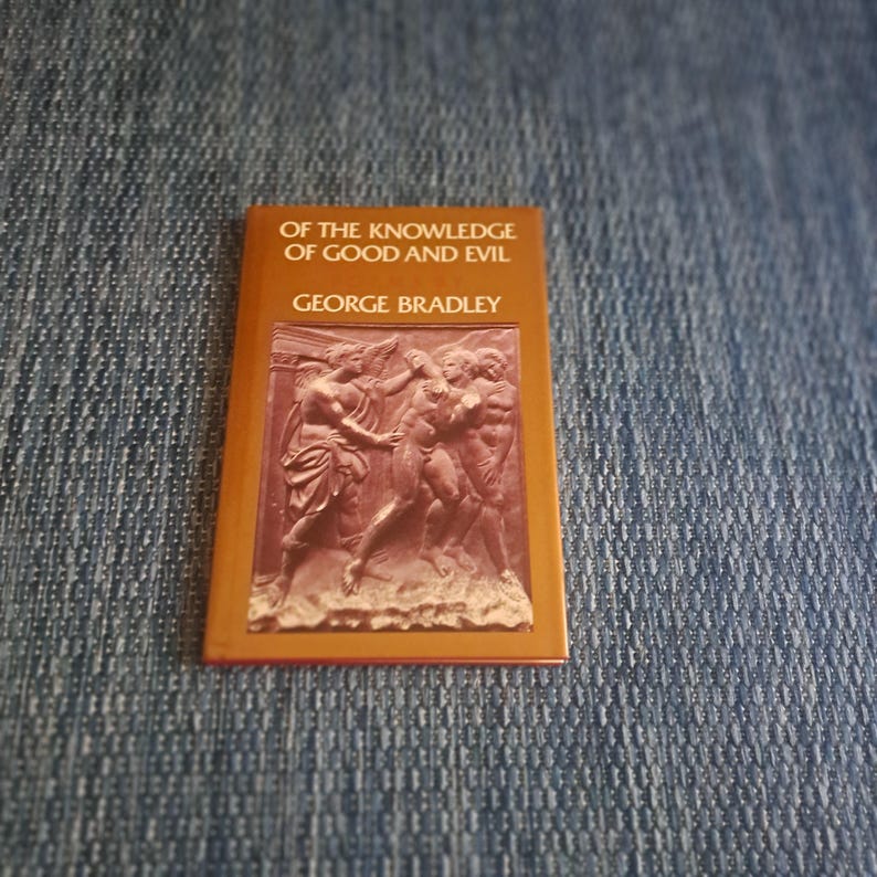 May include: A small, rectangular book with a brown cover featuring the title "OF THE KNOWLEDGE OF GOOD AND EVIL" and the author's name, "GEORGE BRADLEY." The cover also displays a relief sculpture of figures. The book is set against a blue denim background.