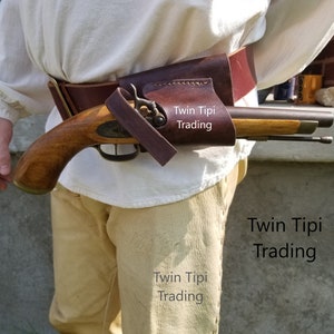 May include: A brown leather holster with a vintage wooden pistol. The pistol has a brass trigger and barrel. The holster is secured to a belt. The text "Twin Tipi Trading" is visible.
