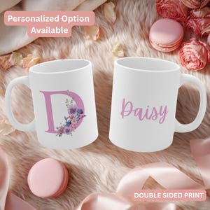 May include: Two white ceramic mugs. One mug features a large pink letter "D" with a floral design. The other mug has the name "Daisy" in pink script. The mugs are on a soft, textured surface.