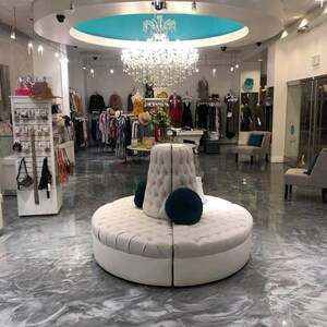 May include: A white, circular, tufted bench with a teal pillow sits in the center of a boutique with a silver epoxy floor. The boutique has a white ceiling with a turquoise dome and a crystal chandelier.