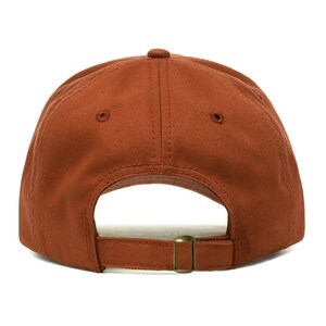 May include: A solid rust-colored baseball cap, viewed from the back. The cap features a curved brim, a fabric strap with a metal buckle for adjustment, and a small button on top. The hat is made of a durable fabric.