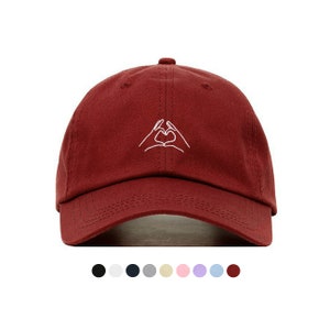 Peut inclure: Une casquette de baseball bordeaux avec un motif brodé blanc de deux mains formant un cœur.