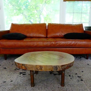 May include: A cognac-colored leather sofa with two black rectangular throw pillows, positioned behind a low coffee table featuring a natural, irregular wood slab top supported by four cylindrical wooden legs.