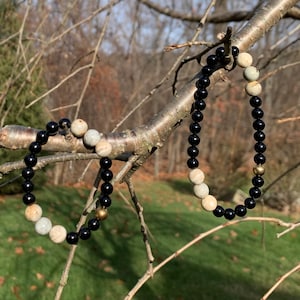 May include: Two bracelets with black and white beads and gold accents. The bracelets are hanging from a tree branch.