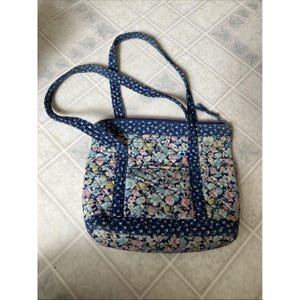 May include: A quilted tote bag with a floral pattern in shades of pink, blue, and green. The bag has a navy blue background and straps. The bag has a front pocket and a zipper closure.