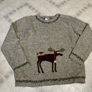 May include: A beige speckled knit sweater featuring a dark brown moose appliqué wearing a red scarf and hat, with a small red bird on its back. The neckline, cuffs, and hem are decorated with a green vine and red berry embroidered pattern.
