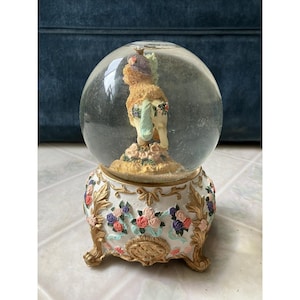 May include: A decorative snow globe featuring a figurine of a horse with a purple hat and floral accents. The base is white with gold trim and colorful flowers. The globe is filled with water and glitter.