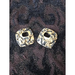 May include: A pair of gold-toned earrings with a unique, abstract design. Each earring features a square shape with a central opening and a textured, swirling pattern in black and gold. The earrings are set against a dark, patterned background.