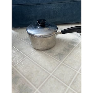 May include: A stainless steel saucepan with a lid and a black handle. The pot is round with a silver finish and a black lid. The handle is black and has a loop at the end. The saucepan is sitting on a tiled surface.