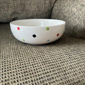 May include: White ceramic bowl with colorful polka dots in red, green, blue, and black. The bowl has a rounded shape and is suitable for serving food. The bowl is sitting on a textured surface.