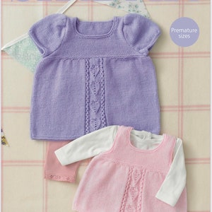 May include: A lilac and pink knitted baby dress set. The lilac dress has short sleeves and a heart-shaped design down the front. The pink dress is sleeveless and layered over a white long-sleeved shirt. The text "SNUGGLY 4 PLY" is at the bottom.