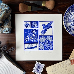 May include: A blue and white linocut print depicting a butterfly, hummingbird, bee, snail, flowers, and a berry. The print is surrounded by a blue and white teacup, postcards, a book, and art supplies on a wooden surface.