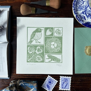 May include: A square linocut print on white paper featuring sage green illustrations of a bird, a strawberry, a ladybug, a bird nest with three eggs, pansy flowers, and a frog. The print includes the handwritten text Spring Garden and a signature.