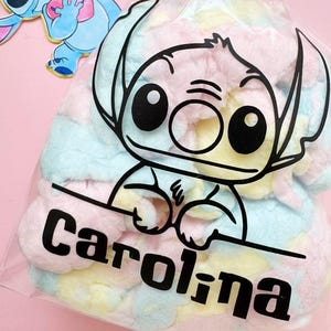 May include: A clear plastic bag filled with pastel-colored treats, featuring a black outline of a cartoon character's face and the name "Carolina" in bold black letters. A cartoon character is visible in the background.