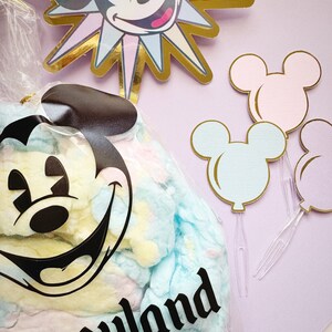 Pu&ograve; includere: Un sacchetto di plastica trasparente pieno di zucchero filato blu pastello, giallo e rosa. Il sacchetto ha una testa di Topolino nera con un grande sorriso e la parola "Disneyland" stampata su di esso. Sullo sfondo ci sono quattro palloncini a forma di testa di Topolino, due rosa, uno blu e uno bianco.