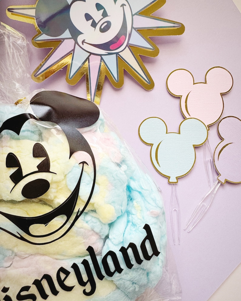 Pu&ograve; includere: Un sacchetto di plastica trasparente pieno di zucchero filato blu pastello, giallo e rosa. Il sacchetto ha una testa di Topolino nera con un grande sorriso e la parola "Disneyland" stampata su di esso. Sullo sfondo ci sono quattro palloncini a forma di testa di Topolino, due rosa, uno blu e uno bianco.