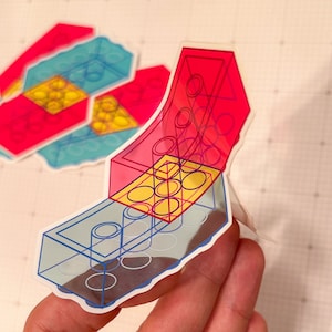 May include: A sticker of a multi-colored Lego brick design. The sticker is transparent and shows the outline of the Lego brick. The sticker is on a hand.