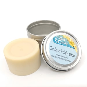 May include: A silver tin with a white lid containing a round, off-white bar of hand salve. The tin has a blue and yellow label that reads "Silver Lining Goods" and "Gardener's Salv-ation ultra moisturizing for hard working hands".