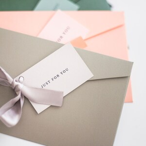 May include: A close-up of several pastel-colored envelopes. A gray envelope in the foreground has a tag that reads "JUST FOR YOU" tied with a light purple ribbon. Other envelopes are in shades of pink, green, and blue.