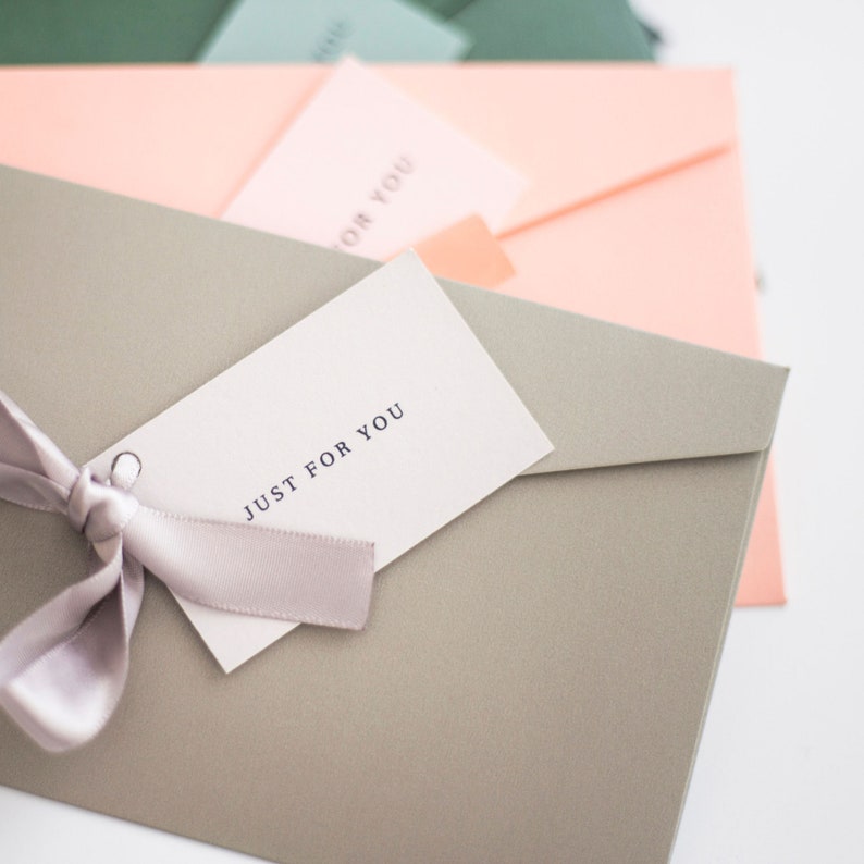 May include: A close-up of several pastel-colored envelopes. A gray envelope in the foreground has a tag that reads "JUST FOR YOU" tied with a light purple ribbon. Other envelopes are in shades of pink, green, and blue.