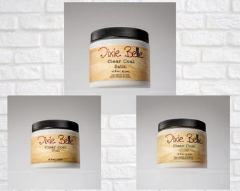 May include: Three white plastic jars with black screw-on lids, each featuring a tan label with the text Dixie Belle Clear Coat. The labels specify the finishes as Satin, Flat, and Gloss, with a volume of 16 fl oz and 473 mL printed on each.