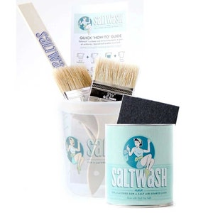 May include: A blue and white can of Saltwash paint for creating a layered sun and salt air soaked look. The can is surrounded by paintbrushes, a sponge, and a wooden stirring stick. The label features a vintage illustration of a woman painting with a brush.